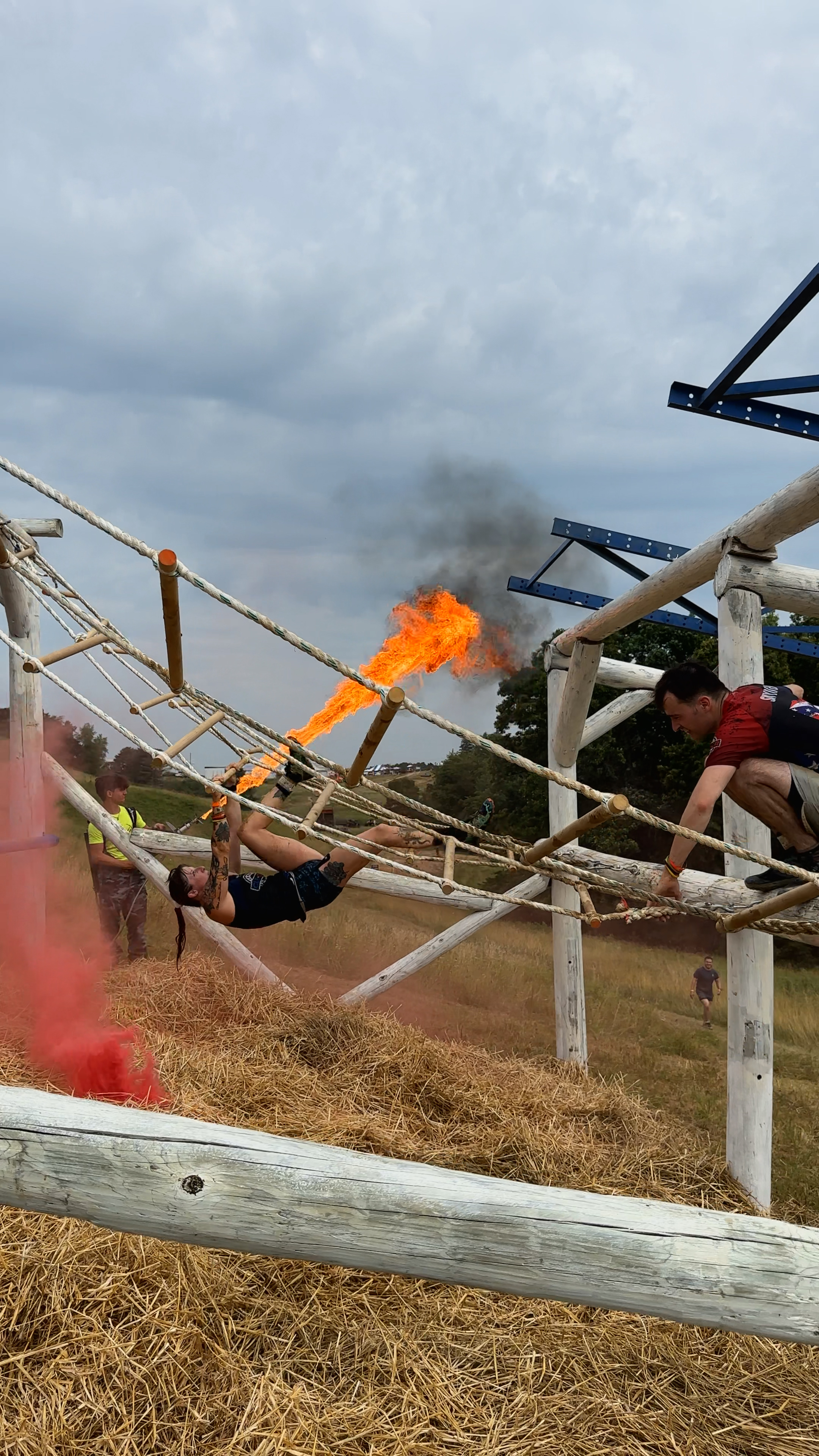 Man in yellow shirt and black shorts holding handles and sliding on an obstacle course with fire behind him.