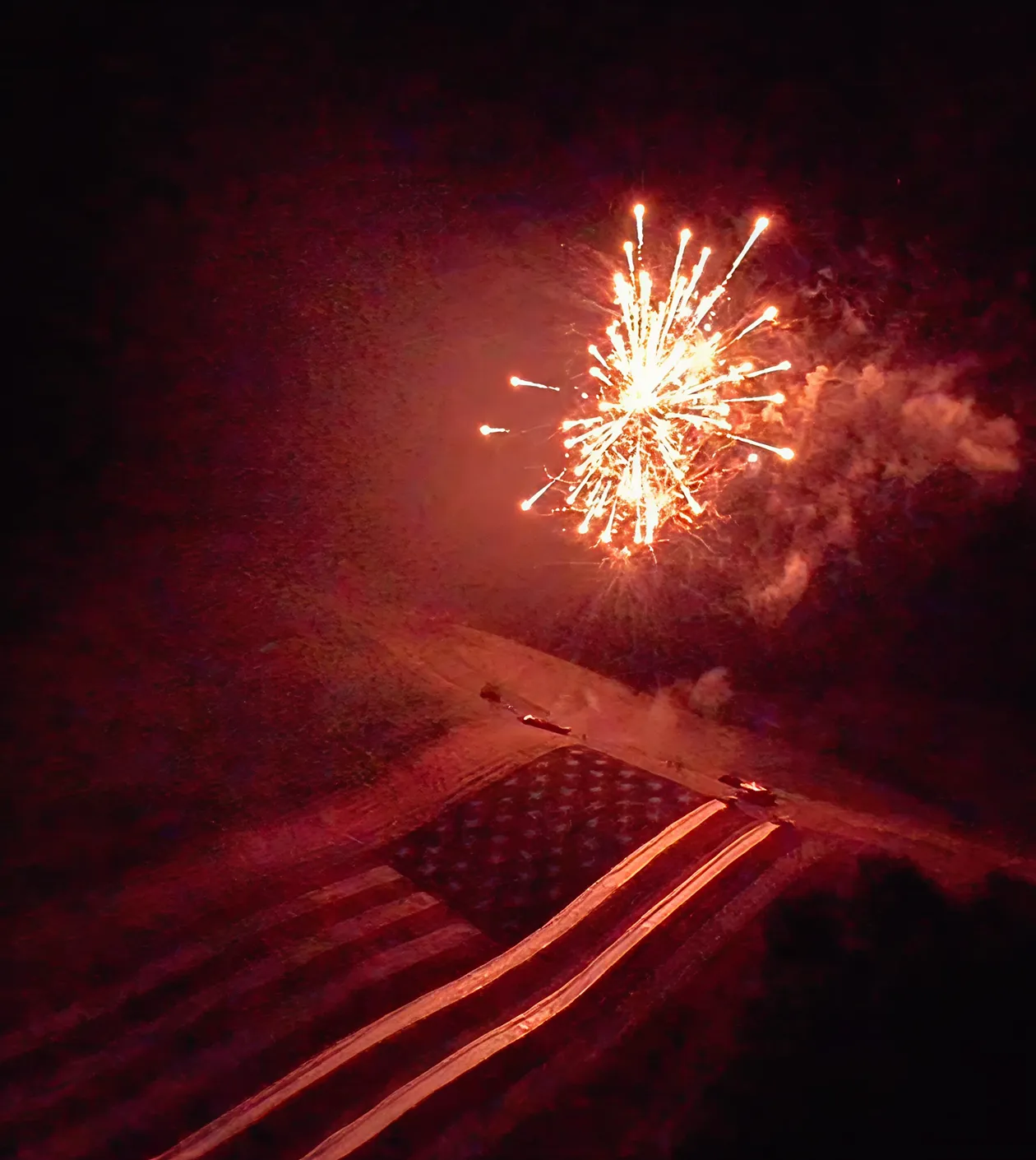 Fireworks explode in the night sky above a large American flag illuminated on the ground.