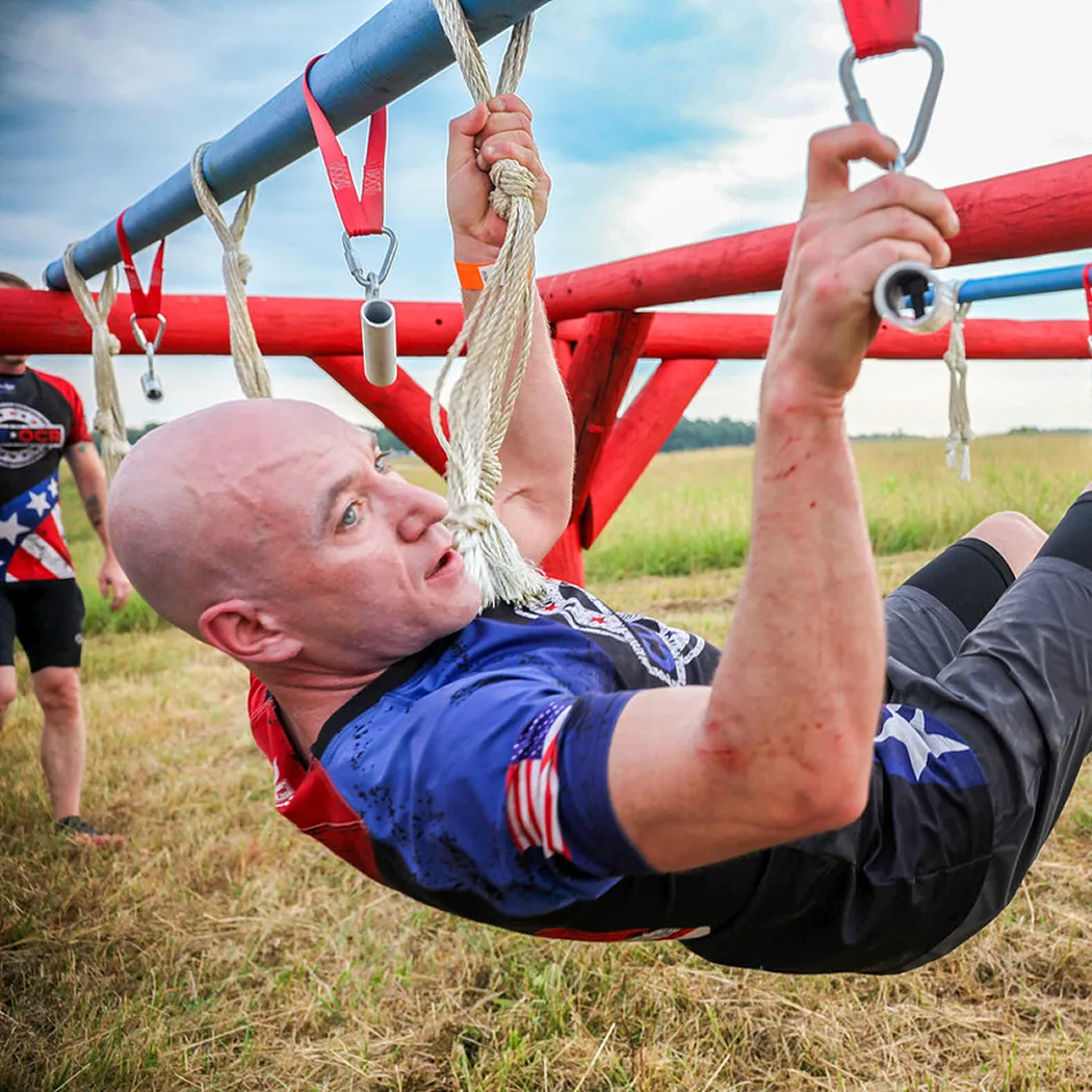Man in athletic gear gripping ropes and metal rings while navigating an outdoor obstacle course with red and blue bars.