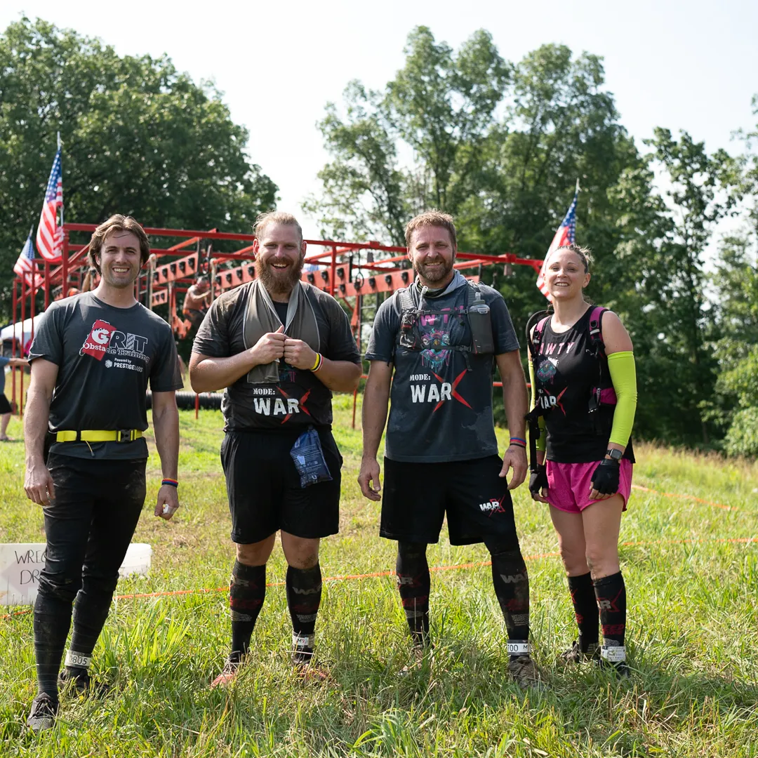 Four adults smiling outdoors in athletic gear with an obstacle course and American flags in the background.