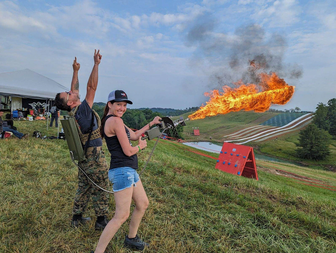 Woman firing a large flamethrower with a man standing behind her raising both hands in excitement on a grassy hillside with an American flag painted on the hill in the background.