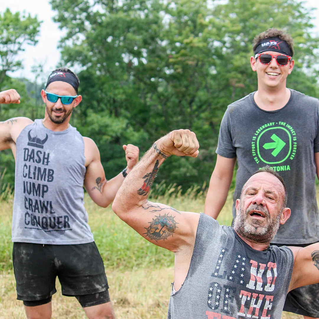 Three men outdoors flexing their arms and smiling, wearing athletic clothing and sunglasses with a green field and trees in the background.