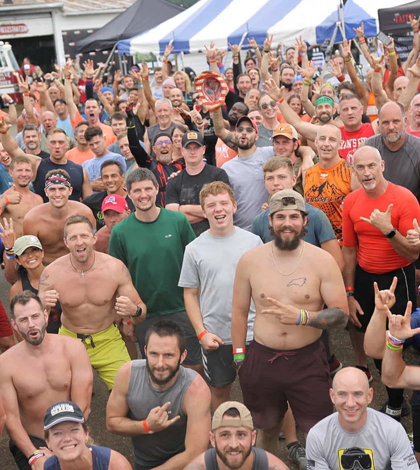 Large group of mostly men outdoors smiling, some shirtless, wearing athletic gear, many making hand gestures.