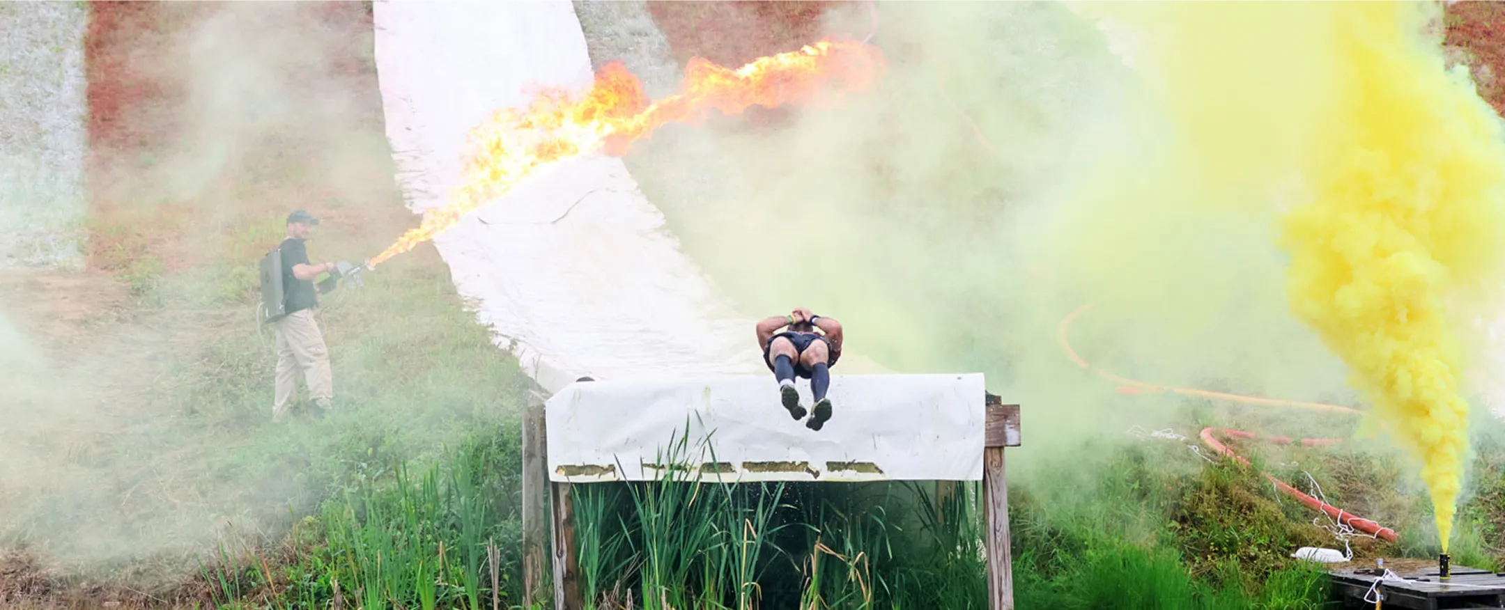 Person sliding down a grass-covered slope over a wooden platform, while another person uses a flamethrower nearby and yellow smoke fills the air.