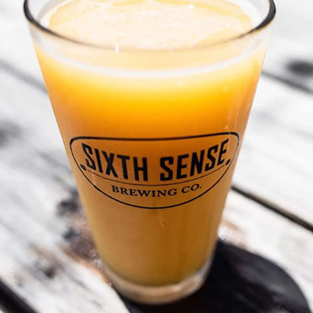 Pint glass with orange-colored beer and foam, labeled 'SIXTH SENSE BREWING CO.' on a wooden table.