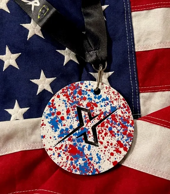 A black medal with red, white, and blue paint splatters and a silver 'X' symbol rests on a rock with an American flag and fireworks in the background.