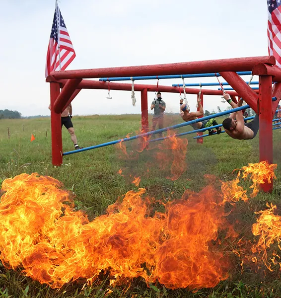 Man in yellow shirt and black shorts holding handles and sliding on an obstacle course with fire behind him.
